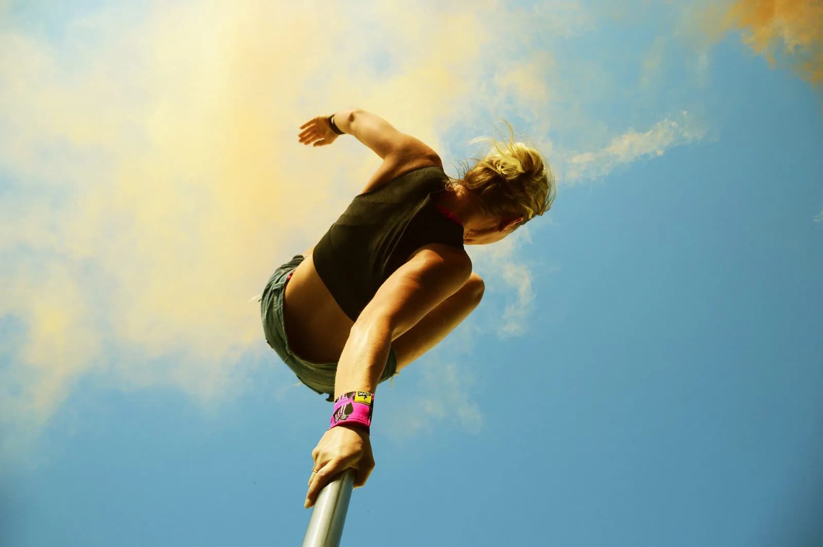 Pole dancer crouched high on an outdoor pole against a blue sky with yellow smoke, holding the pole with one hand and wearing a pink wrist wrap
