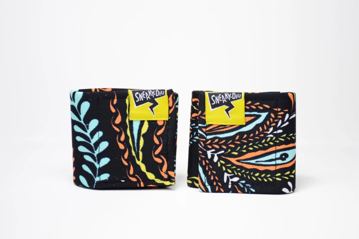 A pair of black wrist wraps with an orange, teal and yellow leaf pattern and yellow SneakyDeez labels, standing on a white background