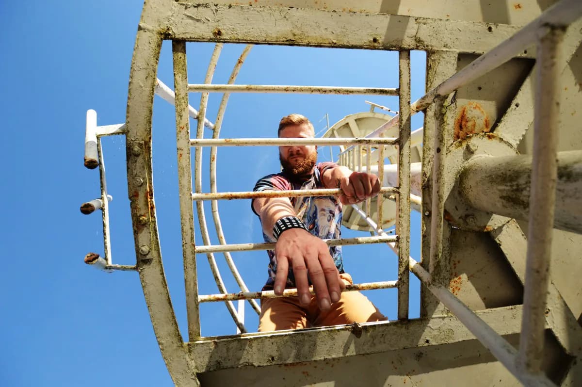 Bearded man seen from below gripping the white bars of a metal tower ladder with both hands against a blue sky, wearing patterned wrist wraps
