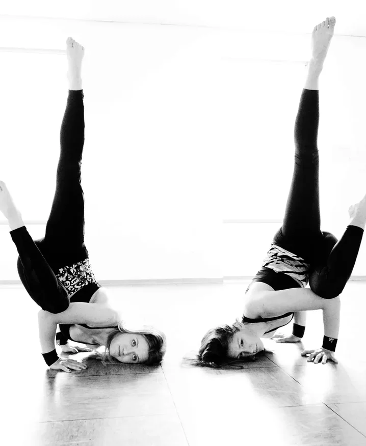Black and white photo of two dancers balancing upside down on their hands with their legs split in the air, both wearing black wrist wraps
