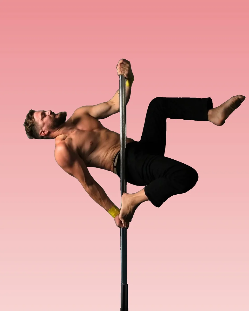 Male pole dancer holding himself sideways off a chrome pole with one hand above his head and the other low on the pole, wearing a yellow wrist wrap